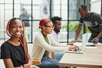 young African American woman in casual clothes posing for the camera while sitting at the table against the background of her colleagues