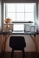 A minimalist home office setup with a modern black desk, a blank open laptop, flower vase, photo frames, and a lamp near a bright window, creating a serene workspace