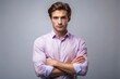 © Achiraya - Confident young man with a strong pose, arms crossed, stands against a light grey background, exuding self-assurance and determination in a light purple shirt.