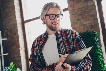 Photo of man wearing checkered stylish casual shirt and eyeglasses writing details clipboard notes information indoors office coworking