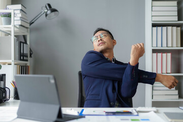 Wall Mural - Young businessman is stretching his arms while sitting at his desk at the office, taking a break from work