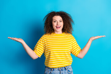 Wall Mural - Photo portrait of pretty young girl compare hold empty space wear trendy striped yellow outfit isolated on blue color background