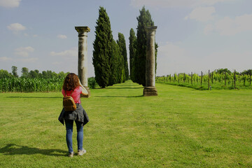 Wall Mural - Woman photographing a majestic cypress avenue with two Roman-style columns in the foreground