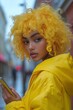 © ArtCookStudio - Young woman with curly yellow hair in a yellow jacket holding a phone on a city street