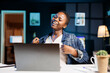 © DC Studio - While sitting at her apartment desk, smiling black woman watches comedy film on digital laptop. Enthusiastic African American lady uses personal computer to communicate with friends virtually online.
