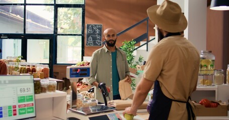 Canvas Print - Man choosing eco products to buy, approaching merchant at register to pay for bag of homegrown produce. Buyer doing grocery shopping at local zero waste eco shop. Handheld shot.