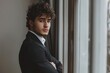 © Olsek - A young man with curly hair stands by a window, dressed in a stylish black suit. He appears thoughtful and introspective. Ideal for business or personal branding. Generative AI