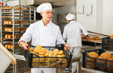 Baker in uniform holding crate with bread, industrial kitchen of bakery on background