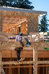 Wall Mural - New residential home construction in framing stage, workman with pneumatic nail gun on exterior side of house working of first floor overhang roof rafters, sunny summer day
