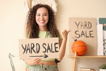 Canvas Print - Young African-American woman holding cardboard with text YARD SALE in room of unwanted stuff