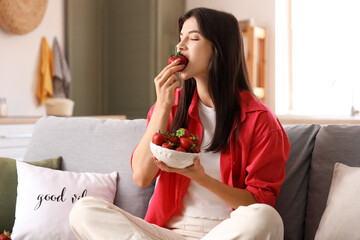 Canvas Print - Beautiful young woman holding bowl with fresh strawberries and sitting on sofa in living room