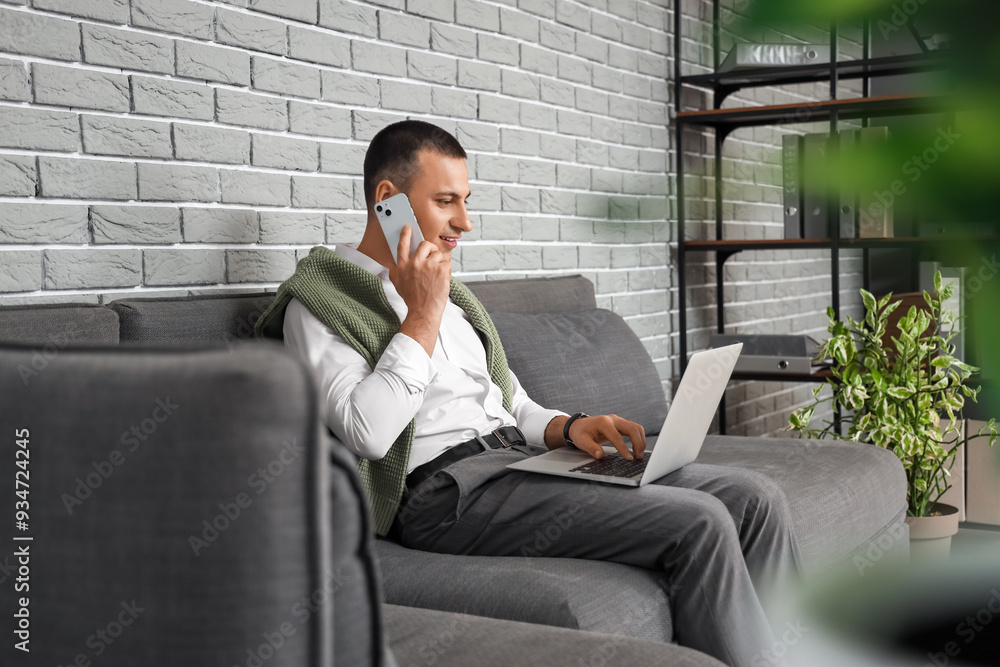 Handsome businessman with laptop talking by mobile phone on sofa in office