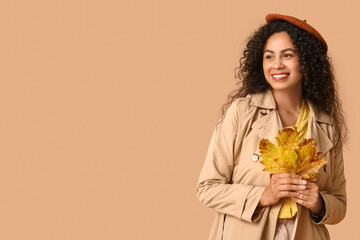 Wall Mural - Beautiful young happy African-American woman in stylish outfit with autumn leaves on beige background