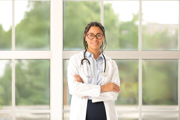 Poster - Young African-American female doctor in eyeglasses with stethoscope near window in clinic