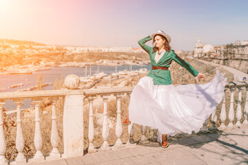 Woman walks around the city, lifestyle. A young beautiful woman in a green jacket, white skirt and hat is sitting on a white fence with balusters overlooking the sea bay and the city.