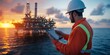 © HendraGalus - Man wearing hard hat and high-visibility jacket holding tablet on ship deck near the ocean with an offshore oil rig at sunset. Maritime technology and industrial work concept