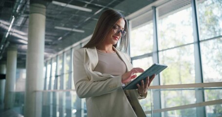 Poster - Happy Female Manager Walking in a Corridor of a Modern Office Building with Glass Walls. Businesswoman Using a Tablet Computer on the Move, Reading a Brief Before Going on a Corporate Meeting