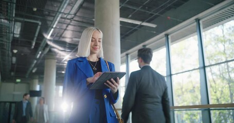 Poster - Young Caucasian Businesswoman Smiling While Checking Financial Documents on a Tablet Computer. Portrait of a Successful Female Corporate Manager Walking in a Spacious Modern Office Building