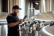 © master1305 - With tablet in hand, brewery technologist checking the brewing machinery, technology control of quality in beer production. Concept of beer, brewery, manufacture, quality control
