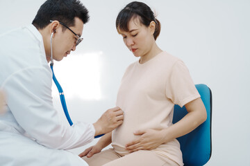 Asian female doctor listens to belly of pregnant mother during a prenatal exam in clinic. doctor provides caring advice, ensuring the health and happiness of the expecting mother and baby.