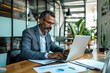 © Ratchpon - Focused businessman working on laptop in modern office, surrounded by plants and documents, conveying sense of concentration and professionalism.
