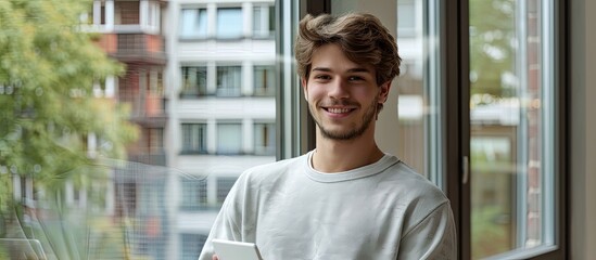 Waist up portrait of an attractive young man holding a digital tablet and smiling at the camera while standing by a window copy space