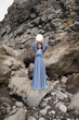 © Milou Dirks - asian young woman in a long blue dress holding round mirror above her head standing near wall of lava rocks in Madeira