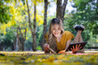 © kamonrat - Smiling Woman Relaxing Outdoors on a Blanket, Reading a Book and Using a Tablet in a Sunlit Park with Yellow Leaves and Greenery