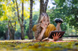 © kamonrat - Smiling Woman Relaxing Outdoors on a Blanket, Reading a Book and Using a Tablet in a Sunlit Park with Yellow Leaves and Greenery
