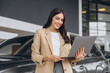 © anatoliycherkas - Portrait of confident successful sales woman using laptop computer standing near new car in car dealership