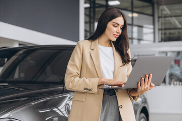 Portrait of confident successful sales woman using laptop computer standing near new car in car dealership