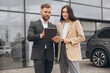 © anatoliycherkas - Bearded business man car salesman in grey suit giving explanations on tablet to pretty young woman in beige jacket outside car dealership