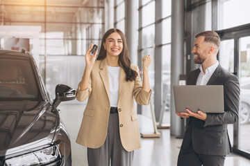 Wall Mural - Charming young Caucasian woman holding keys of new car and show fist and say yes in modern car dealership. Happy girl buyer rejoices after making her dream come true.