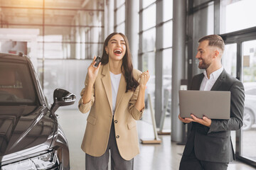 Wall Mural - Charming young Caucasian woman holding keys of new car and show fist and say yes in modern car dealership. Happy girl buyer rejoices after making her dream come true.