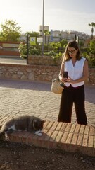 Wall Mural - Woman outdoors checking phone next to a resting cat on a sunny day with urban background