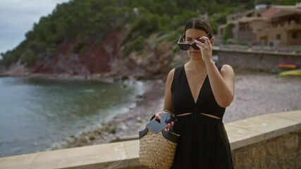 Poster - Young hispanic woman in sunglasses holding a phone and basket by the sea in port de valldemossa, mallorca, spain.