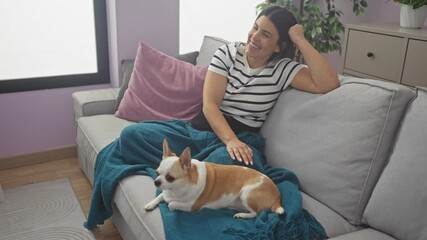 Canvas Print - A young hispanic woman relaxes on a living room couch with her chihuahua dog, laughing joyfully and enjoying a cozy indoor moment at home.