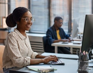 Medium portrait of young female African American computer programmer smiling typing on keyboard while sitting at desk working in IT development office, copy space