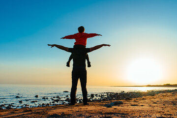 Canvas Print - Father and son walking on lake shore on sunset light, son is sitting on father's shoulder