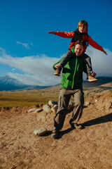 Canvas Print - Father and son tourists walking on high mountains, son is sitting on father's shoulder