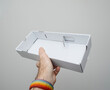 © ifeelstock - A hand holds a shallow, white cardboard box with a simple construction packaging design, set against a neutral gray background, emphasizing minimalist presentation