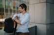 © qunica.com - Professional young man in casual attire organizing documents in his backpack outside a modern office building. Represents business, organization, and efficiency in an urban environment.