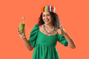 Canvas Print - Beautiful young happy African-American woman with glass of kiwi smoothie showing thumb-up gesture on orange background