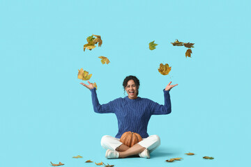 Canvas Print - Beautiful young happy African-American woman in stylish outfit with autumn leaves and pumpkin sitting on blue background