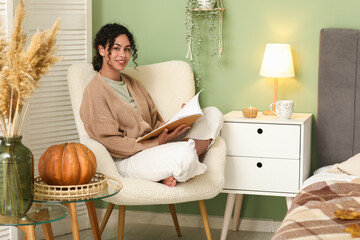 Poster - Beautiful young happy African-American woman in autumn outfit with pumpkin reading magazine on armchair at home