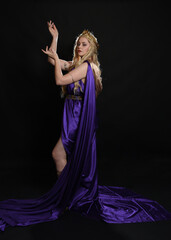 Canvas Print - full length portrait of beautiful blonde female model wearing purple and gold silken toga robes with royal crown, historical fantasy goddess costume. Isolated on dark studio background.