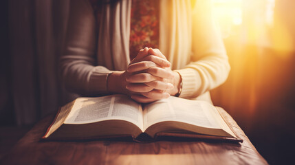 Close-up of a person praying with hands clasped over an open Bible on a wooden table. Spiritual reflection and devotion in a sunlit room symbolizing faith and devotion.