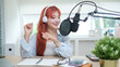 © wattana - Smiling young woman speaking into a professional microphone while recording a podcast in a bright and modern workspace.