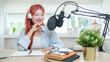 © wattana - Smiling young woman speaking into a professional microphone while recording a podcast in a bright and modern workspace.