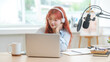 © wattana - Cute young woman with red hair using laptop while recording a podcast in a modern studio.